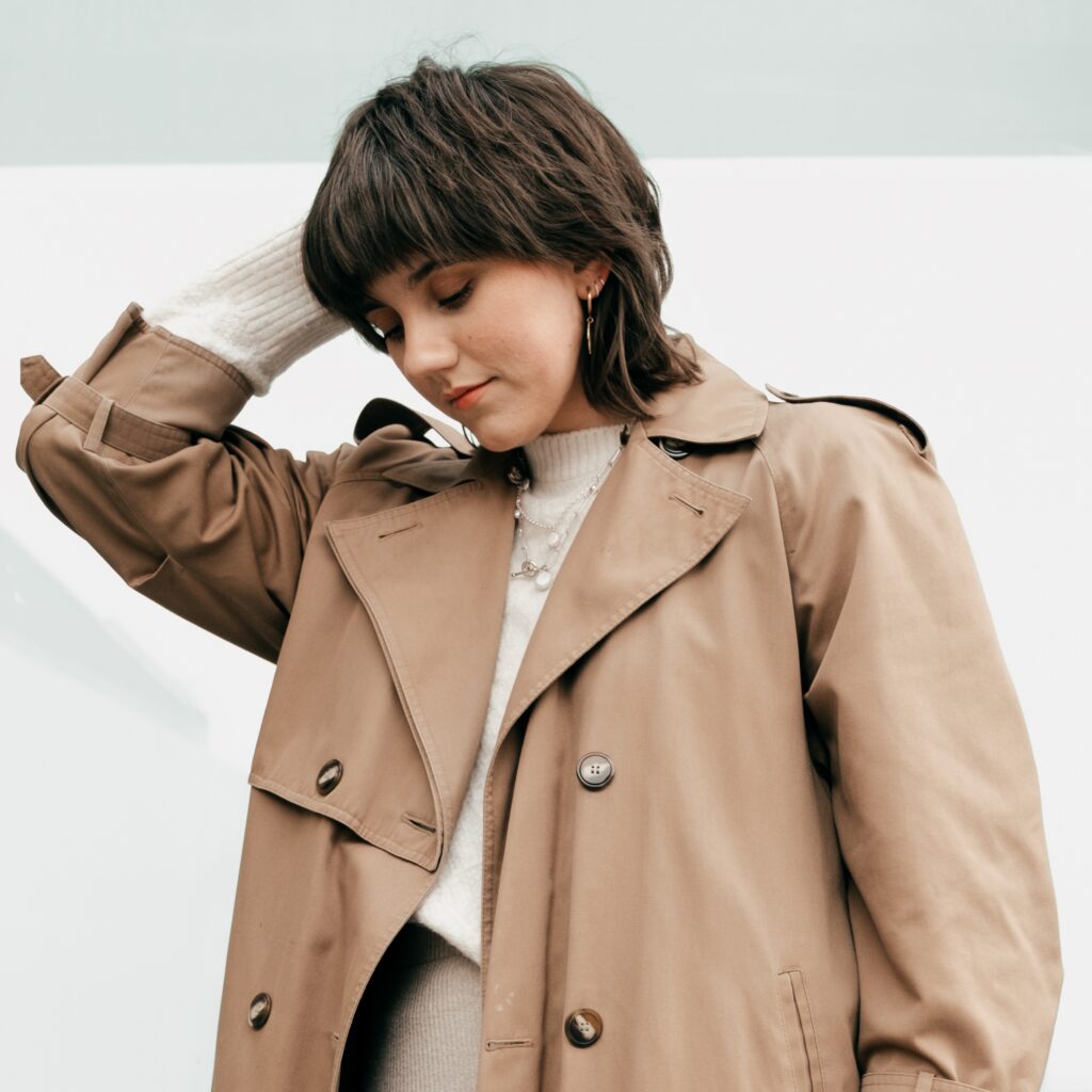 Mujer joven con cabello corto y chaqueta color Mocha Mousse, mirando hacia abajo con un fondo claro, representando los colores tendencia de 2025. A young woman with short hair and a Mocha Mousse-colored coat, looking down against a light background, representing the trend colors of 2025. Colores tendencia de 2025. Trend colors of 2025.