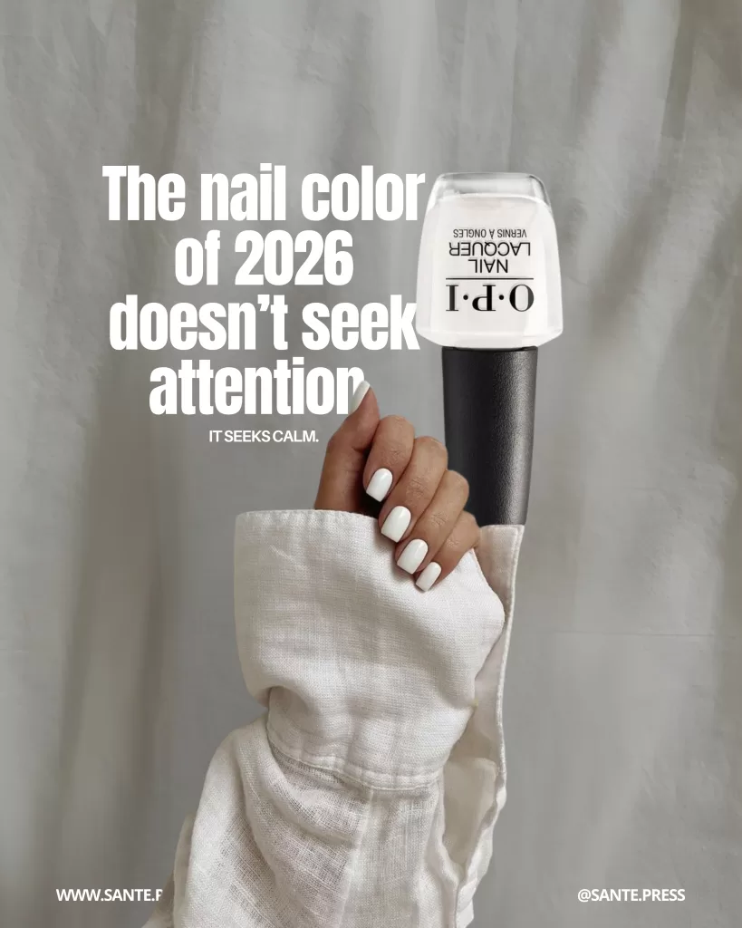 Hand with a white manicure holding an OPI nail polish bottle in front of white fabric; text states that the nail color of 2026 doesn’t seek attention — it seeks calm.
