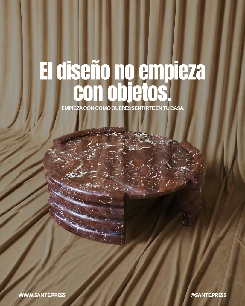 Mesa redonda de mármol rojizo con vetas blancas frente a una cortina beige drapeada, creando una escena cálida y minimalista. Texto: “El diseño no empieza con objetos. Empieza con cómo querés sentirte en tu casa”.