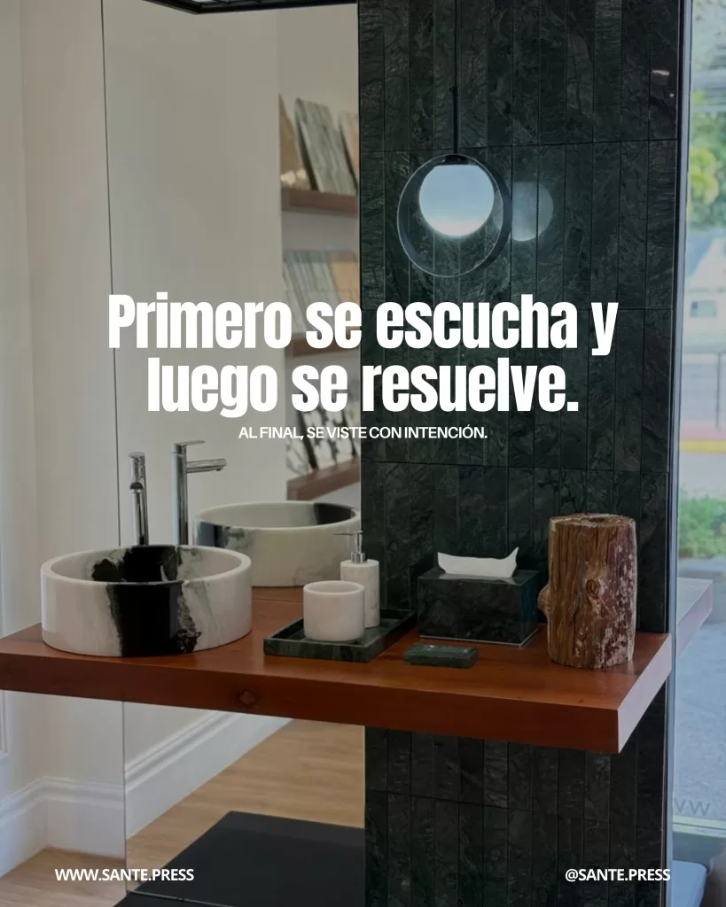 Baño contemporáneo con lavabos de piedra, accesorios de mármol y pared oscura texturizada iluminada por una lámpara circular. Texto: “Primero se escucha y luego se resuelve. Al final, se viste con intención”.