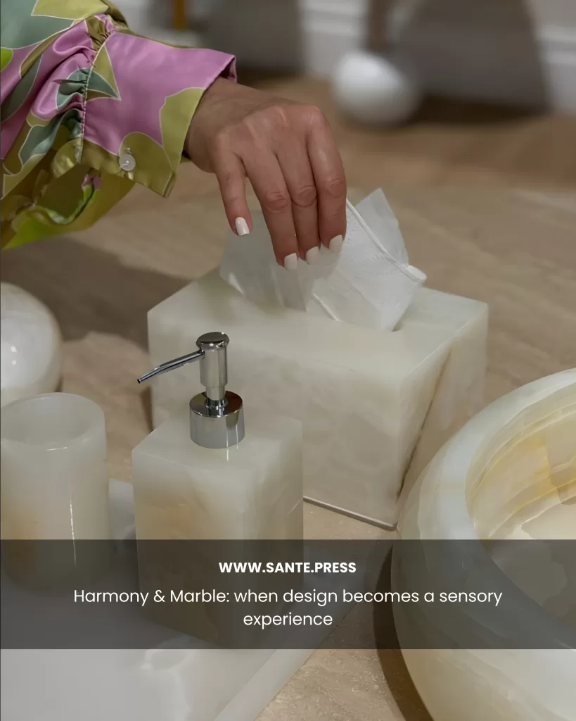 Hand with white nails pulling a tissue from a translucent marble dispenser next to matching bathroom accessories. Text: “Harmony & Marble: when design becomes a sensory experience.”