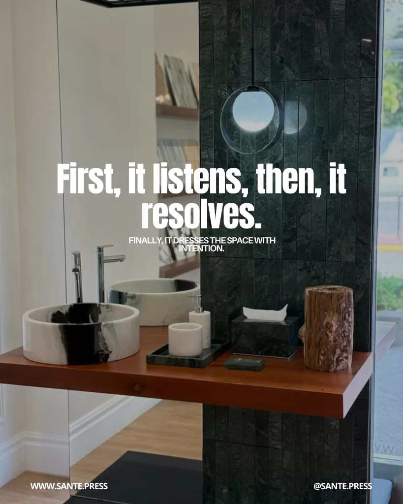 Contemporary bathroom with stone sinks, marble accessories, and a textured dark wall illuminated by a circular lamp. Text: “First, it listens, then it resolves. Finally, it dresses the space with intention.”