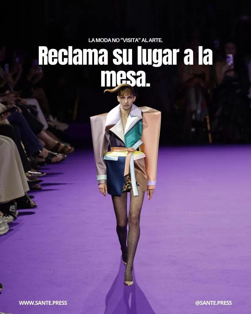 Modelo caminando en pasarela con un conjunto estructural multicolor de silueta arquitectónica sobre una alfombra púrpura, con público a ambos lados. Texto: “La moda no ‘visita’ al arte. Reclama su lugar a la mesa”.