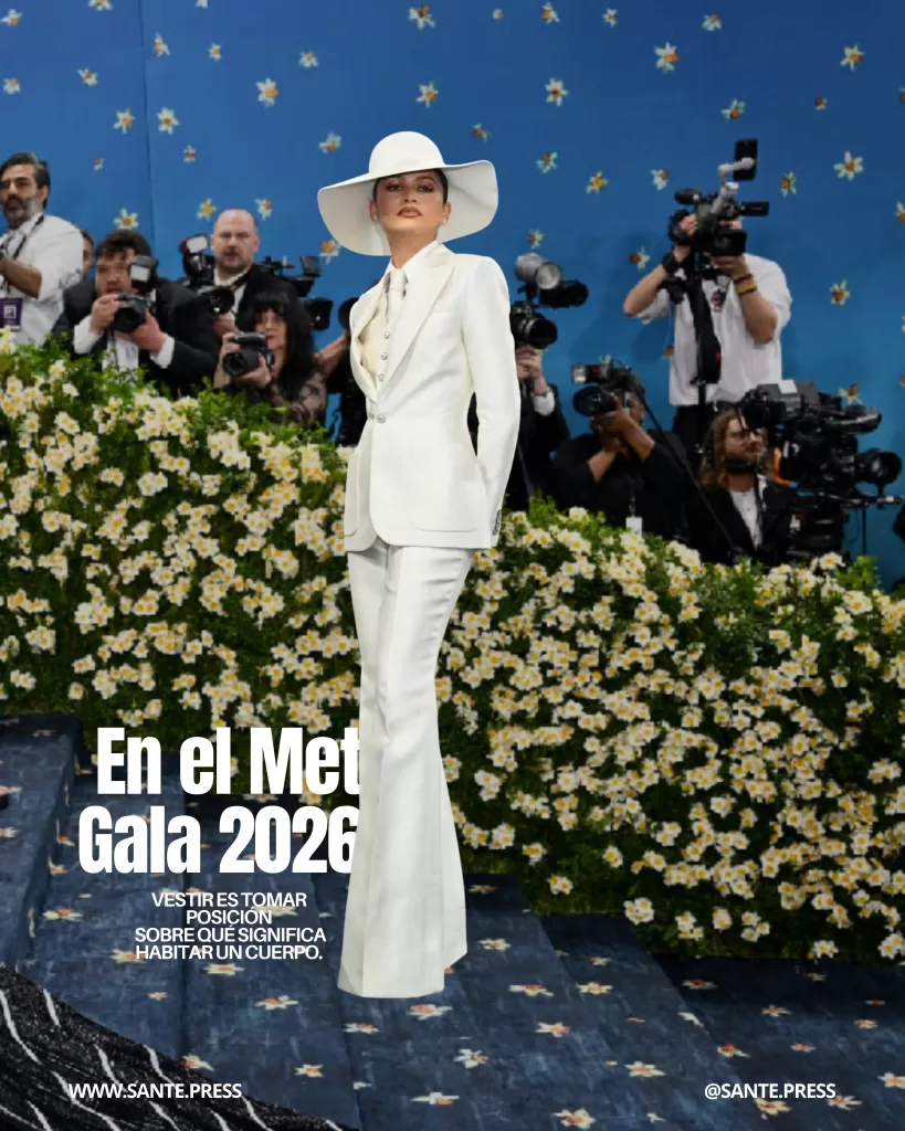 Celebridad en traje blanco impecable y sombrero de ala ancha posando frente a fotógrafos en la alfombra roja del Met Gala, con fondo floral. Texto: “En el Met Gala 2026 vestir es tomar posición sobre qué significa habitar un cuerpo”.