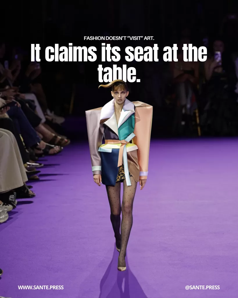 Model walking the runway in a multicolored structural ensemble with an architectural silhouette on a purple carpet, with an audience on both sides. Text: “Fashion doesn’t ‘visit’ art. It claims its seat at the table.”